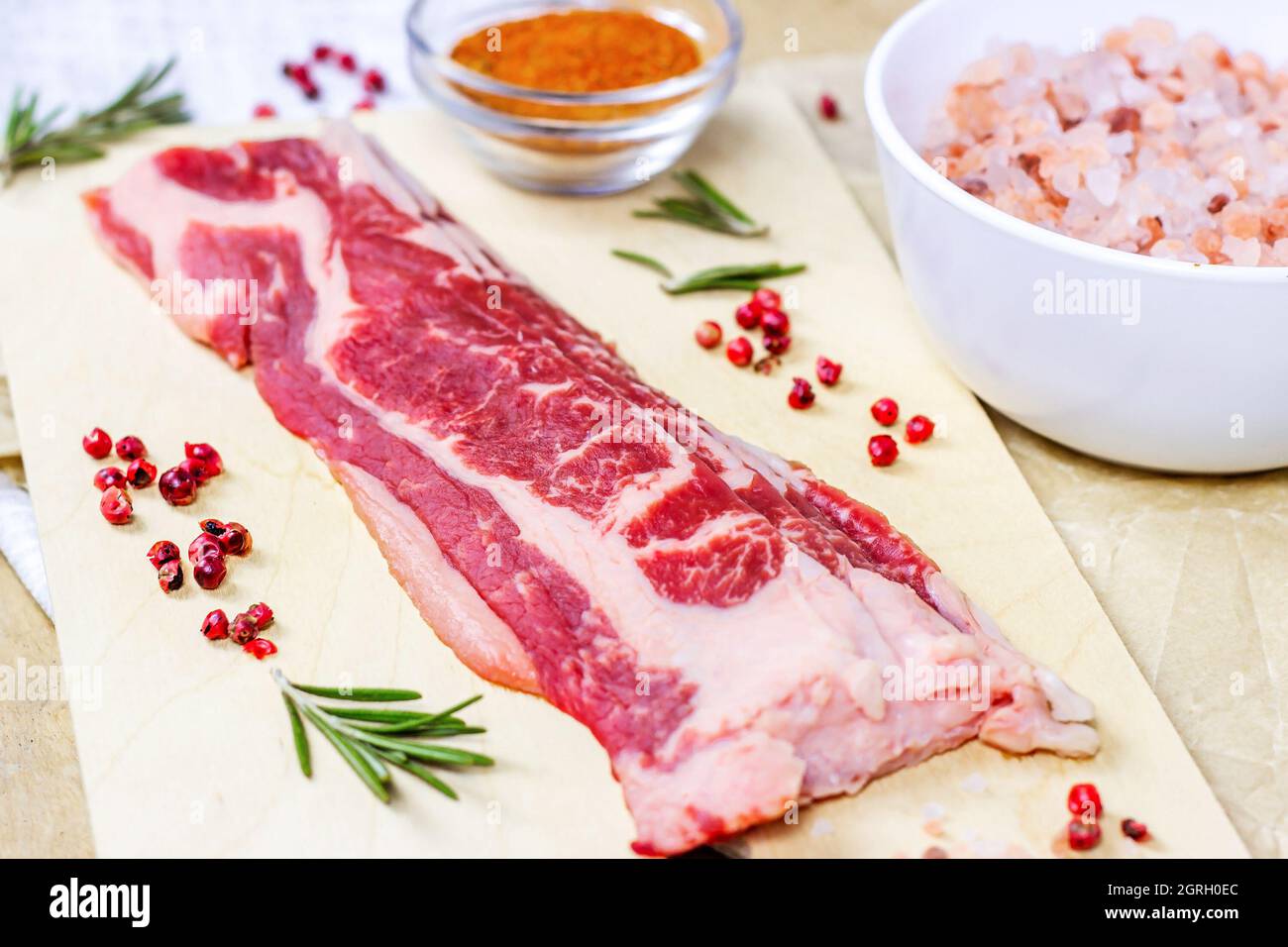 Vista dall'alto di sottili fette di manzo marmorizzato con spezie, rosmarino e sale su tagliere di legno chiaro pronto per la cottura. Foto Stock