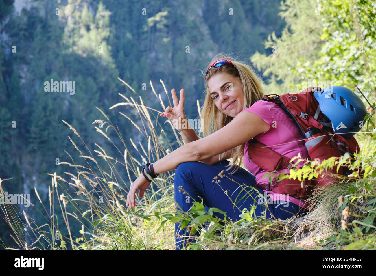 Profilo laterale di donna alpinista con casco attaccato allo zaino, seduto in erba con la mano sollevata come segno di vittoria. Ritratto, avventura. Foto Stock