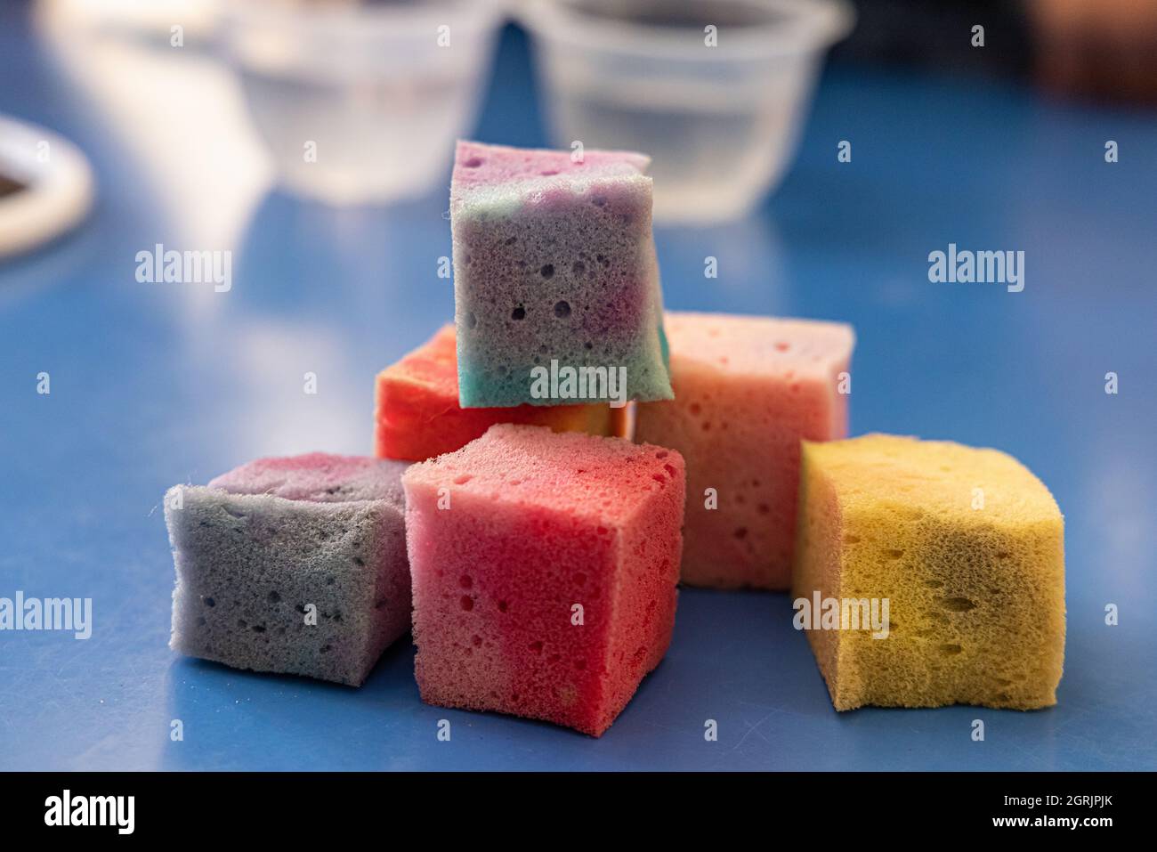 Una spugna tagliata a cubetti quadrati, immersa in vernice di colore diverso, posta su una tavola blu per asciugare, oltre alle ciotole di cristallo riempite d'acqua. Foto Stock