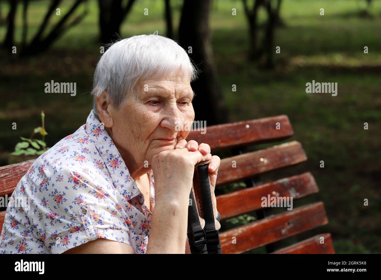 Donna anziana con espressione pacifica del viso seduta con bastoni da passeggio su una panca nel parco. Vita in vecchiaia, concetto di pensionamento Foto Stock