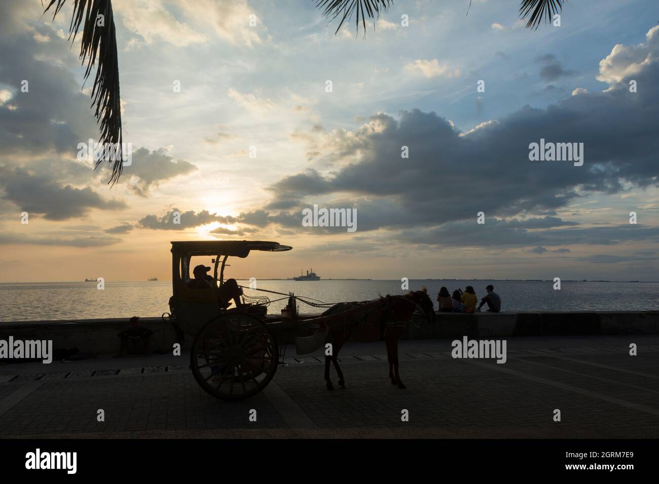 Autista di una carrozza trainata da cavalli a due ruote (kalesa) e alcuni giovani che si godono il tramonto sulla Baywalk a Manila, Filippine. Foto Stock