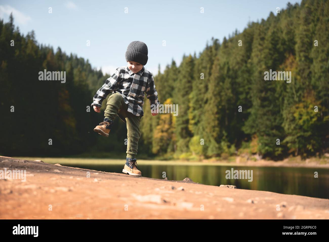 Bambino piccolo in una camicia a quadri e cappello grigio sulla costa del lago forestale. Infanzia con natura concetto di amore Foto Stock