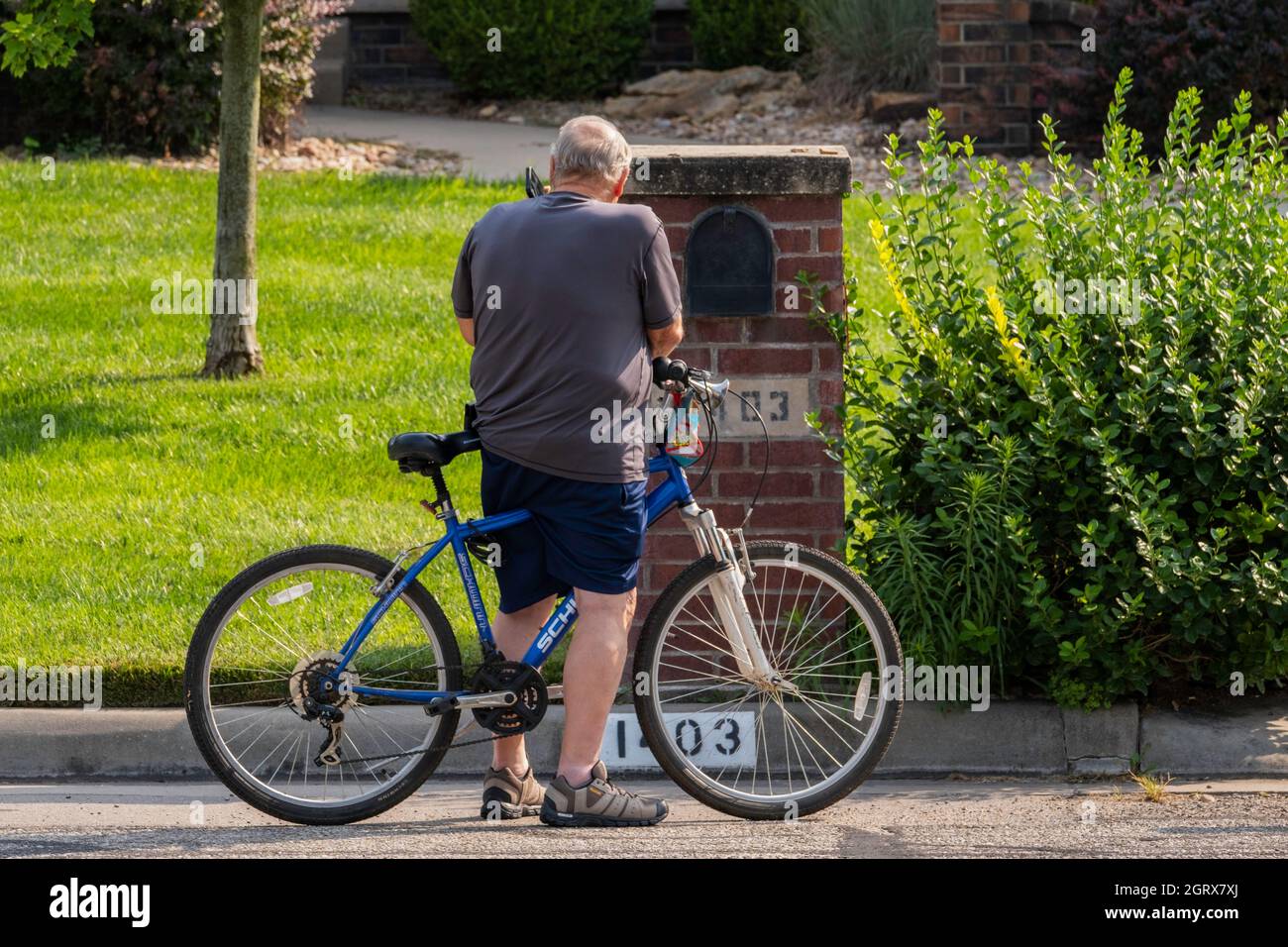 Un uomo caucasico anziano ferma la sua bicicletta per strada per rispondere al suo cellulare in un quartiere residenziale. Kansas, Stati Uniti. Foto Stock