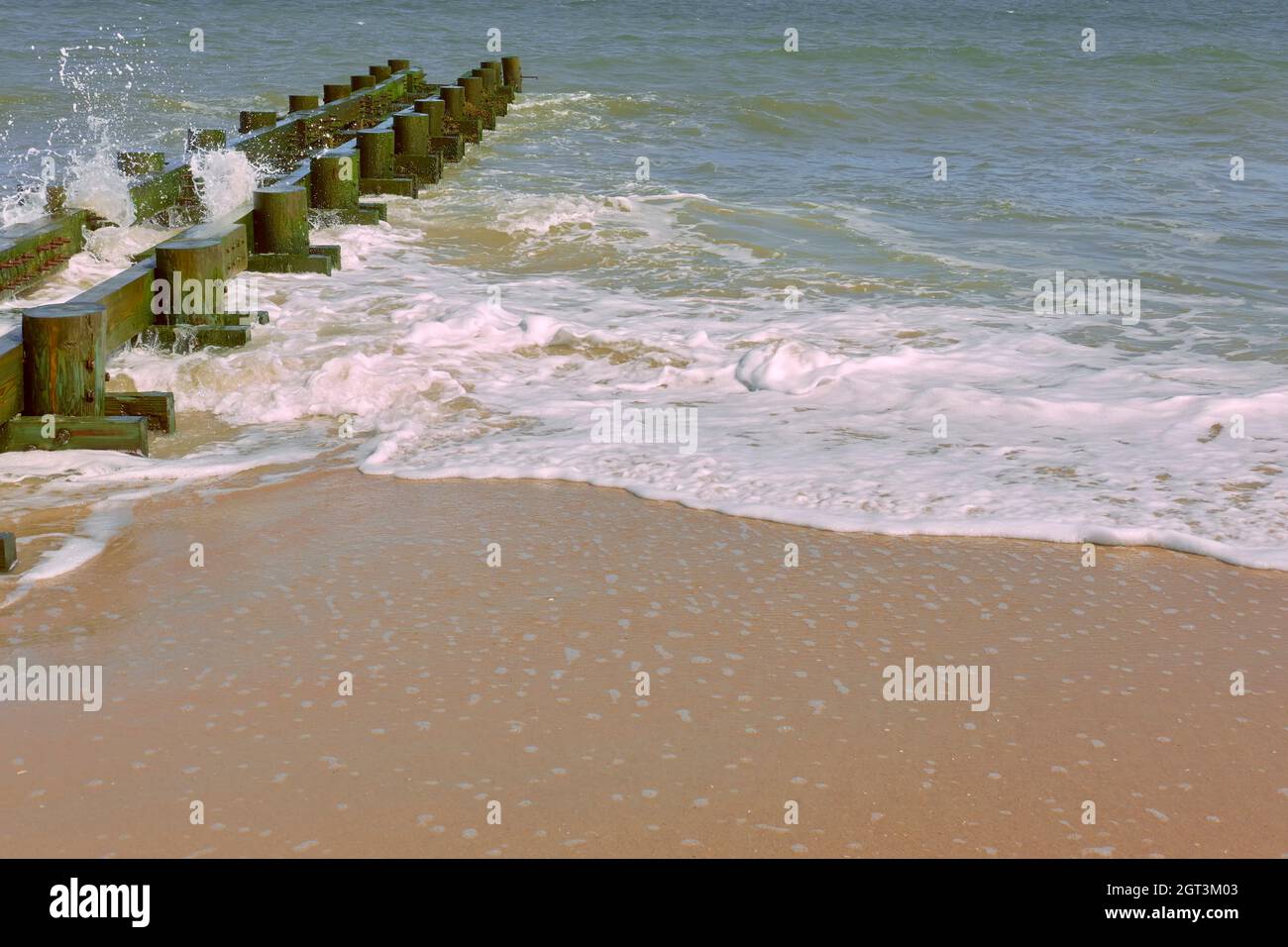 Il surf sull'Oceano Atlantico schiumoso passa davanti a un frangiflutti di legno durante una giornata di sole. Foto Stock