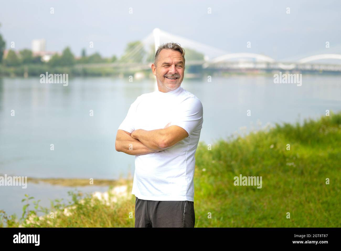 Uomo anziano in piedi su una riva del fiume con braccia ripiegate che guardano da parte con un sorriso raggiante e divertente espressione in una giornata di primavera soleggiata Foto Stock