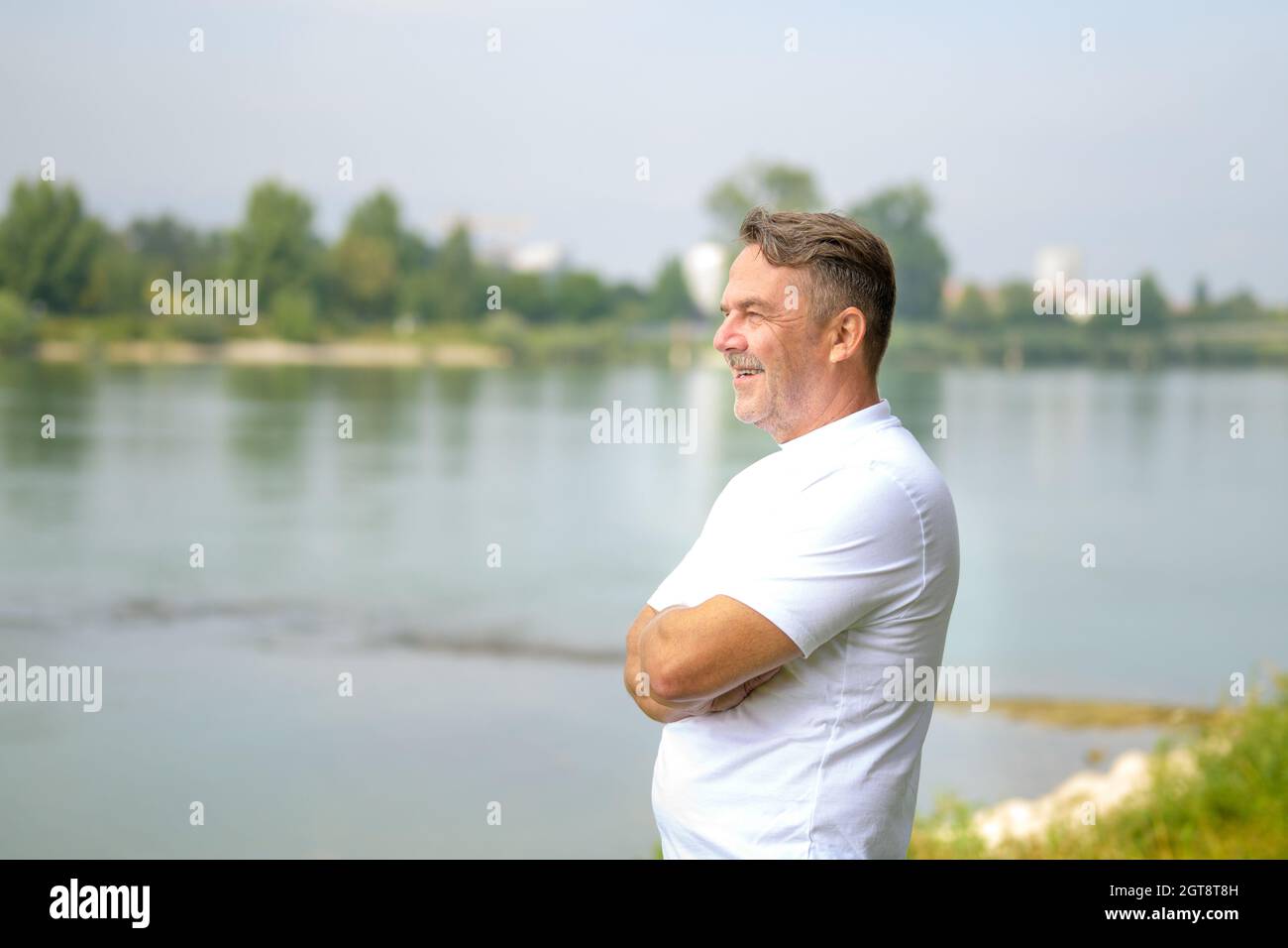 Uomo anziano in piedi su una riva del fiume con braccia ripiegate che guardano da parte con un sorriso raggiante e divertente espressione in una giornata di primavera soleggiata Foto Stock