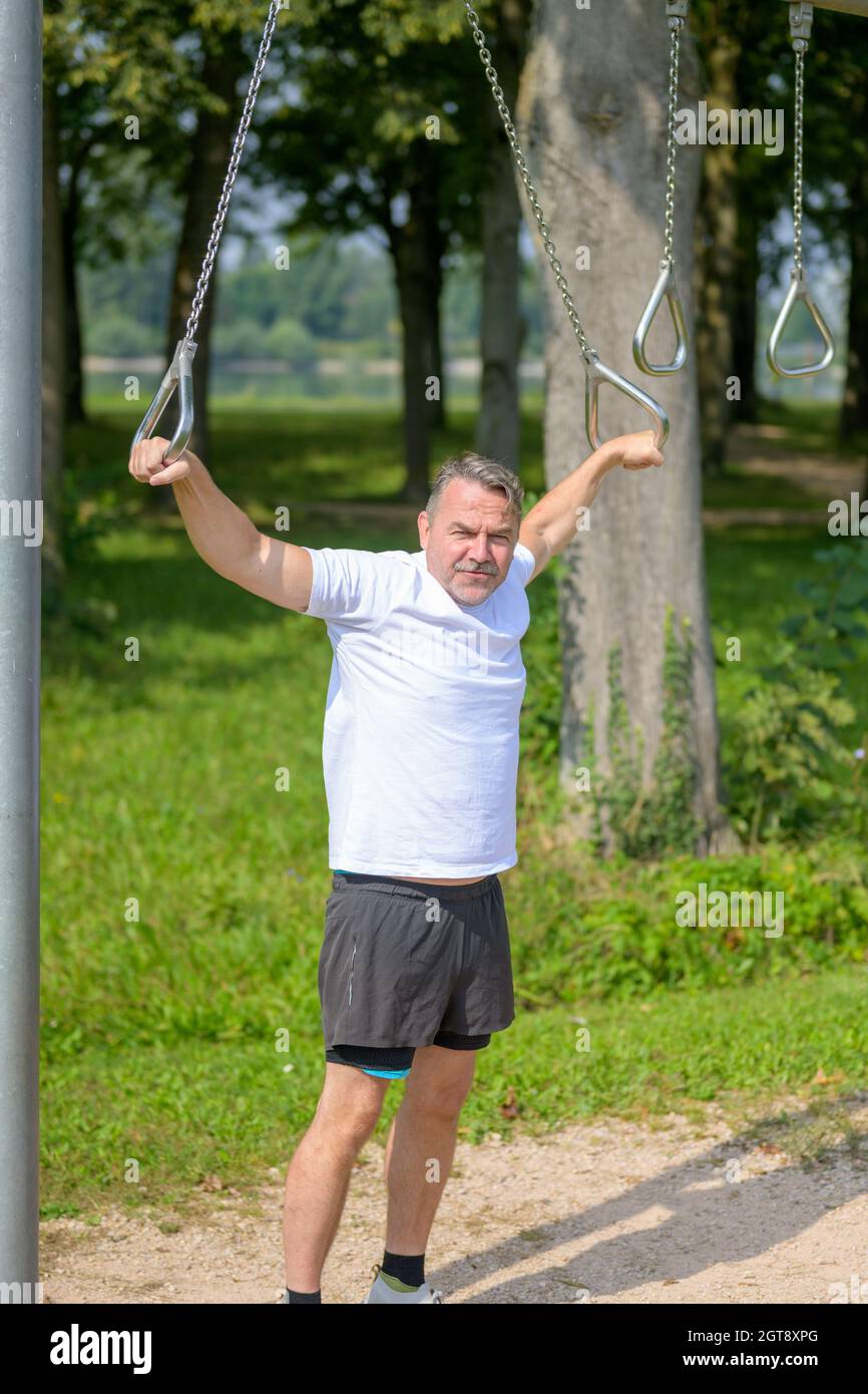 Uomo anziano che utilizza anelli per allenarsi in una struttura sportiva all'aperto in un lussureggiante parco verde in piedi guardando la macchina fotografica con un sorriso in una salute e in forma Foto Stock