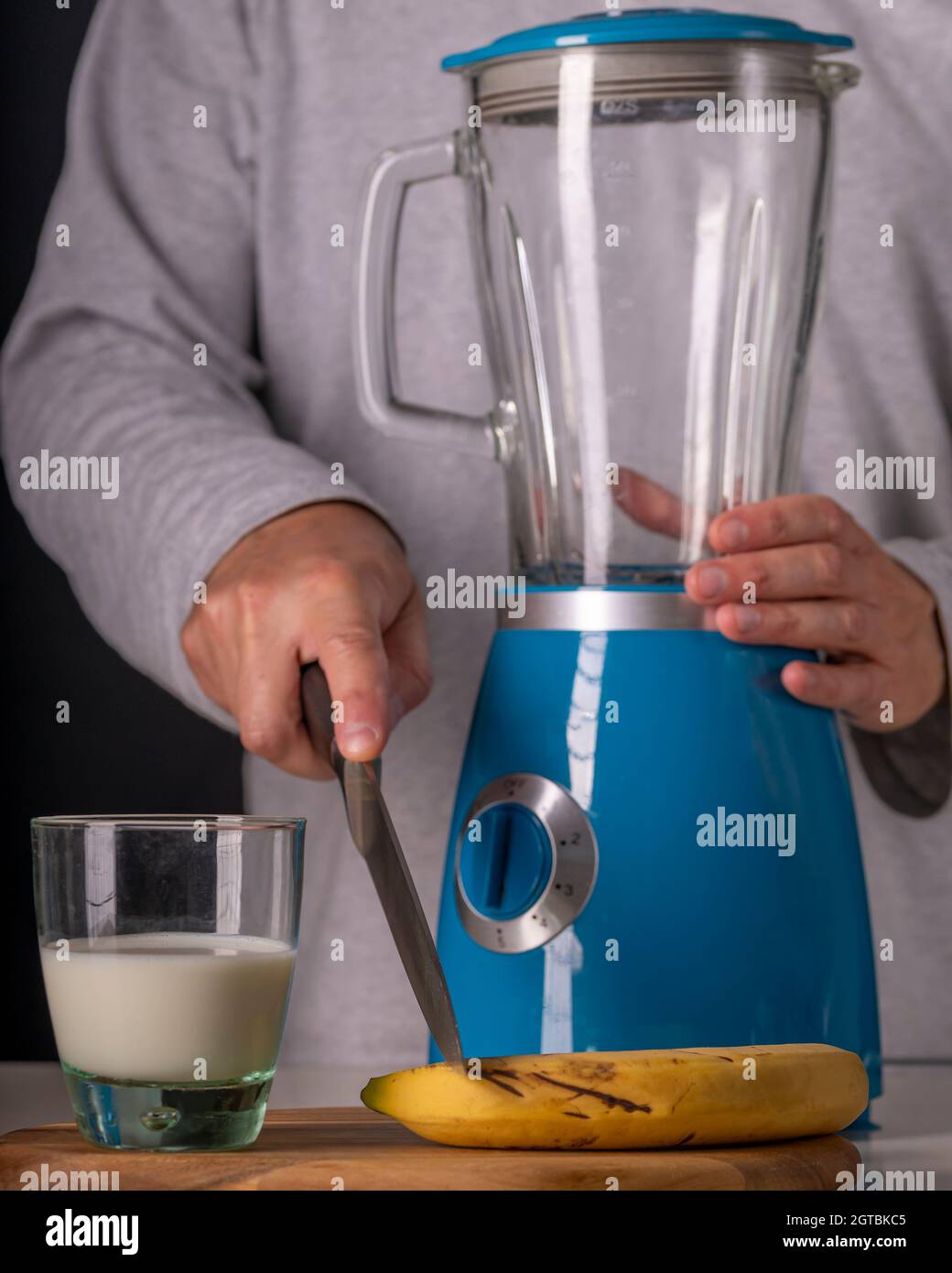Un uomo taglia una banana con un frullatore e un bicchiere di latte nelle vicinanze per preparare un frullato Foto Stock