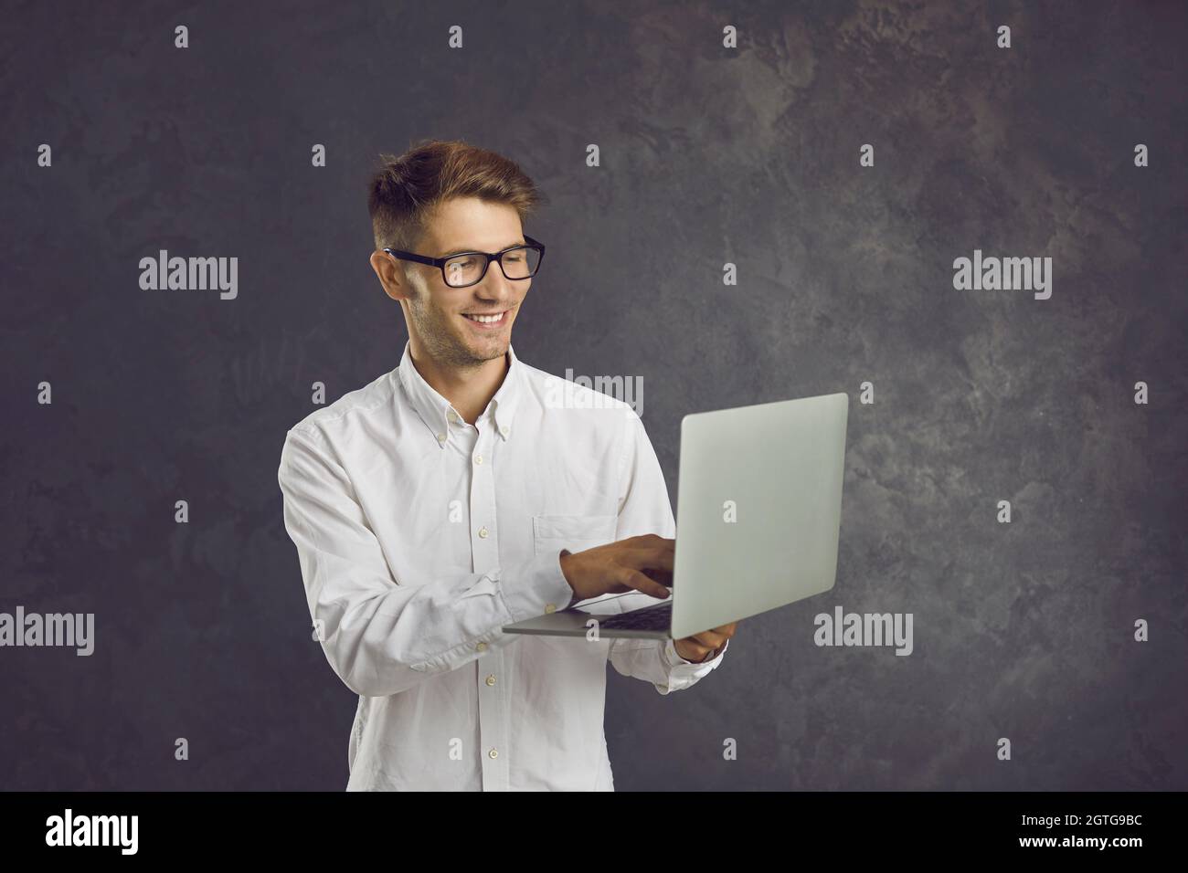 L'uomo sorridente si alza su sfondo grigio e digita il testo sul portatile che tiene nelle mani. Foto Stock