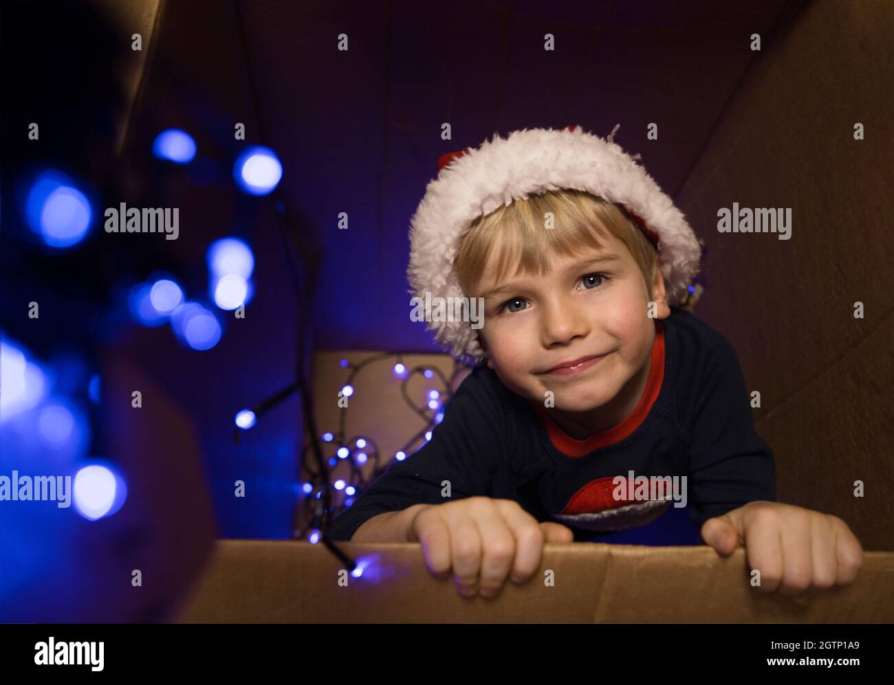 Ragazzo, di 4-5 anni, si siede e guarda fuori da una grande scatola di cartone, fuoco selettivo, rumore, oscurità. Atmosfera natalizia. Bokeh festivo. Divertente Foto Stock