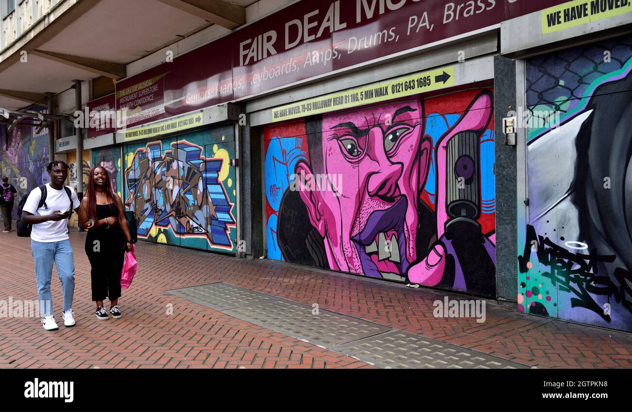 Due persone che camminano lungo il marciapiede, negozi chiusi con arte da parete su persiane Foto Stock