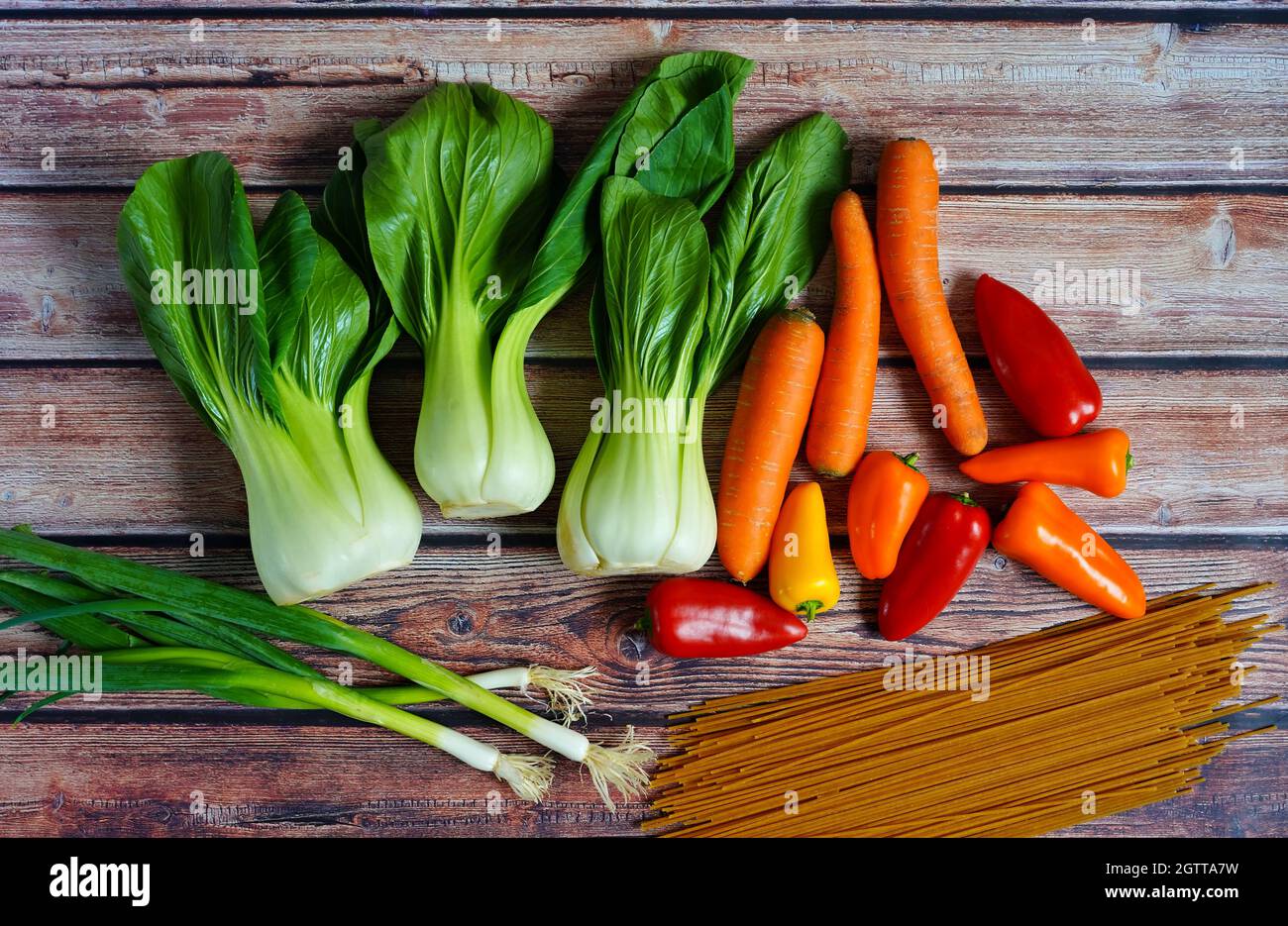 Cipolline fresche, pak choi, carote, peperone e tagliatelle integrali su un rustico tavolo in legno. Preparazione per una cucina casalinga sana. Foto Stock