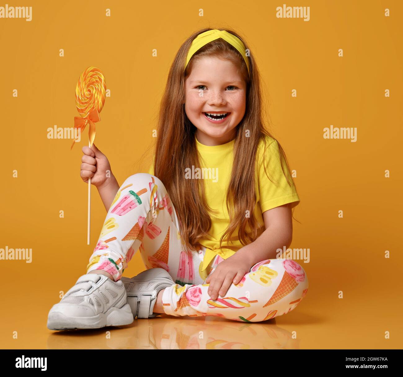 Carina bambina in t-shirt gialla e leggings colorati si siede sul pavimento  tenendo grandi lecca in mano e sorrisi Foto stock - Alamy