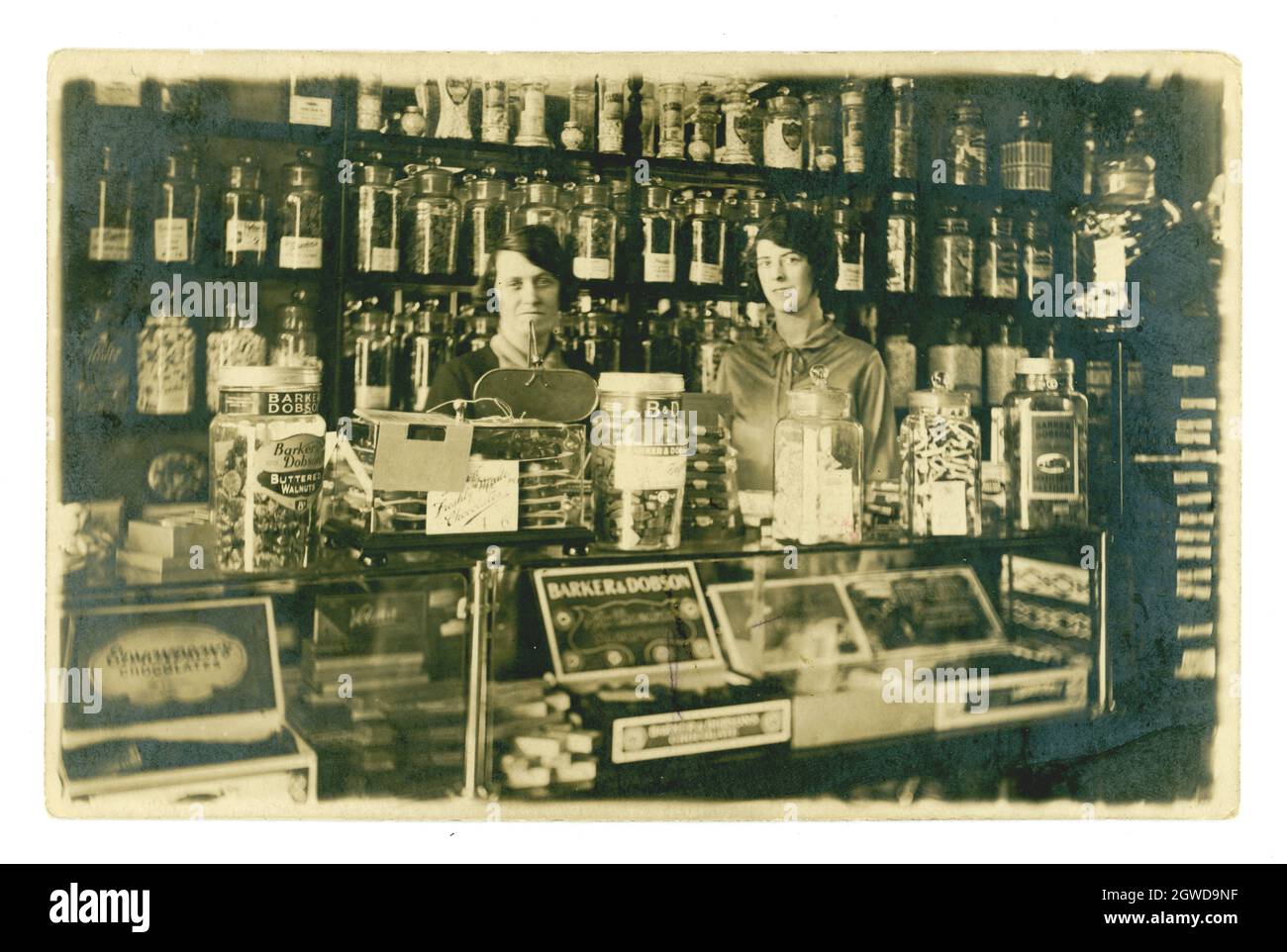 Originale e rara cartolina degli anni '1920 con interni tipici e tradizionali, in vecchio stile, con proprietari o assistenti felici, sorridenti dietro il bancone, il famoso marchio di pasticceria Barker e Dobson su vasetti di vetro. Negozio vittoriano, interno negozio di dolciumi. Di W Williams, Caernarvon, Galles, Regno Unito circa 1927 Foto Stock