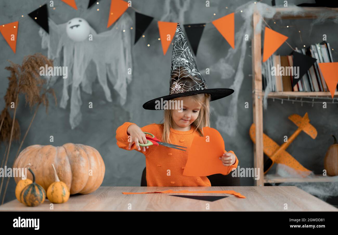 Ragazza caucasica in un costume da strega siede al tavolo a casa e prepara artigianato per la vacanza di Halloween. Foto Stock