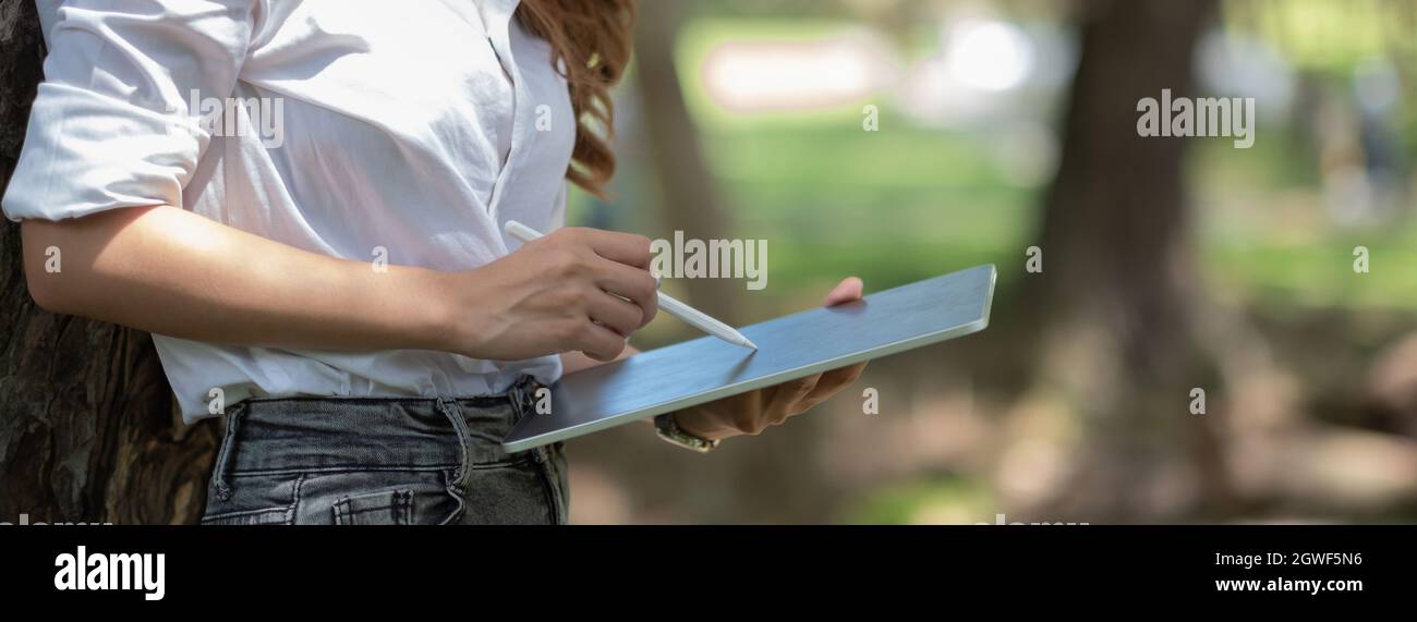 Primo piano mano di donna con penna stilo scrivere su blocco note digitale, toccando lo schermo del tablet digitale lavorando in parco Foto Stock