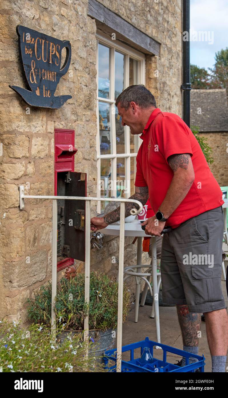 Guiting Power, Gloucestershire, Inghilterra, Regno Unito. 2021. L'operaio postale nella sua camicia rossa uniforme che svuota una cassetta delle lettere nel villaggio rurale di Cotswolds di Guit Foto Stock