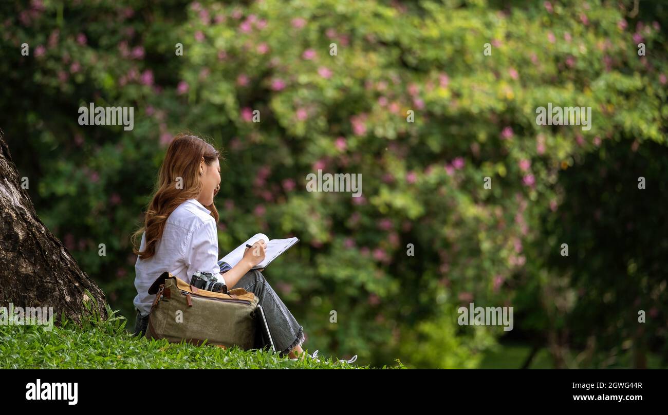 Immagine di una donna gioiosa che tiene diario libro di scrittura nota mentre si siede su erba nel parco. Foto Stock
