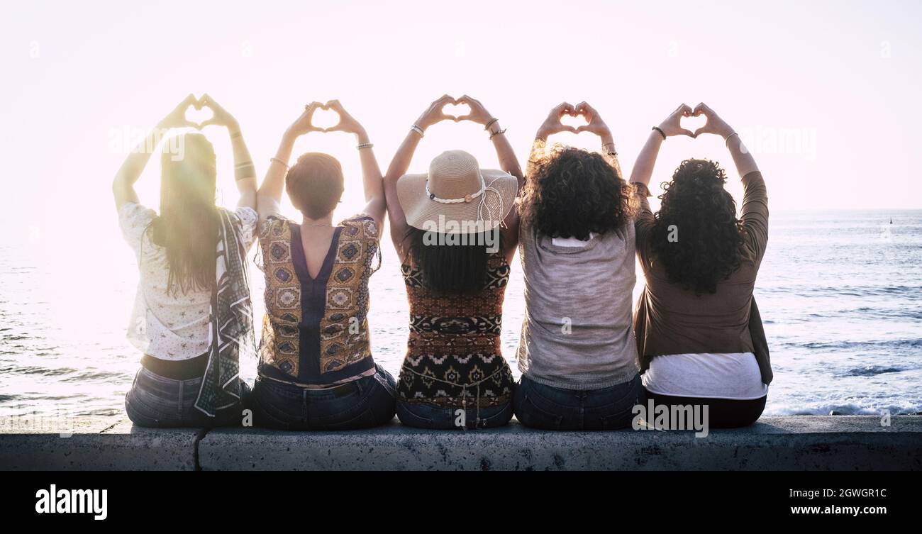 Vista posteriore di cinque donne che si siedono in fila sul muro di mare gesturando l'amore facendo il simbolo della forma del cuore con le mani e ammirando il bel mare. Retro di f Foto Stock