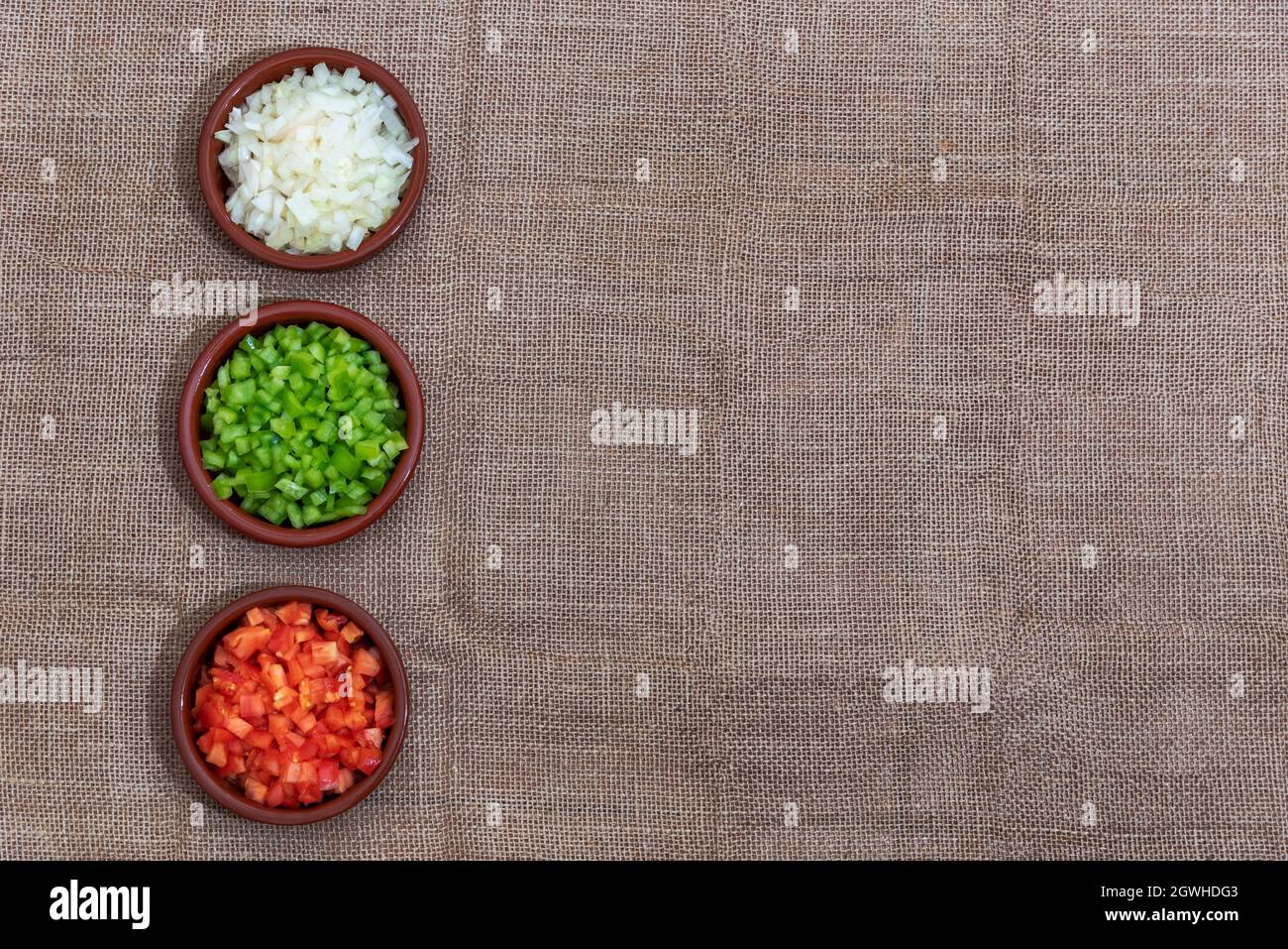 Fondo di tessuto ruvido e di colore marrone, con tre piccoli piatti di argilla con gli ingredienti della salsa, cipolla, pepe e pomodoro.spazio copia. Foto Stock