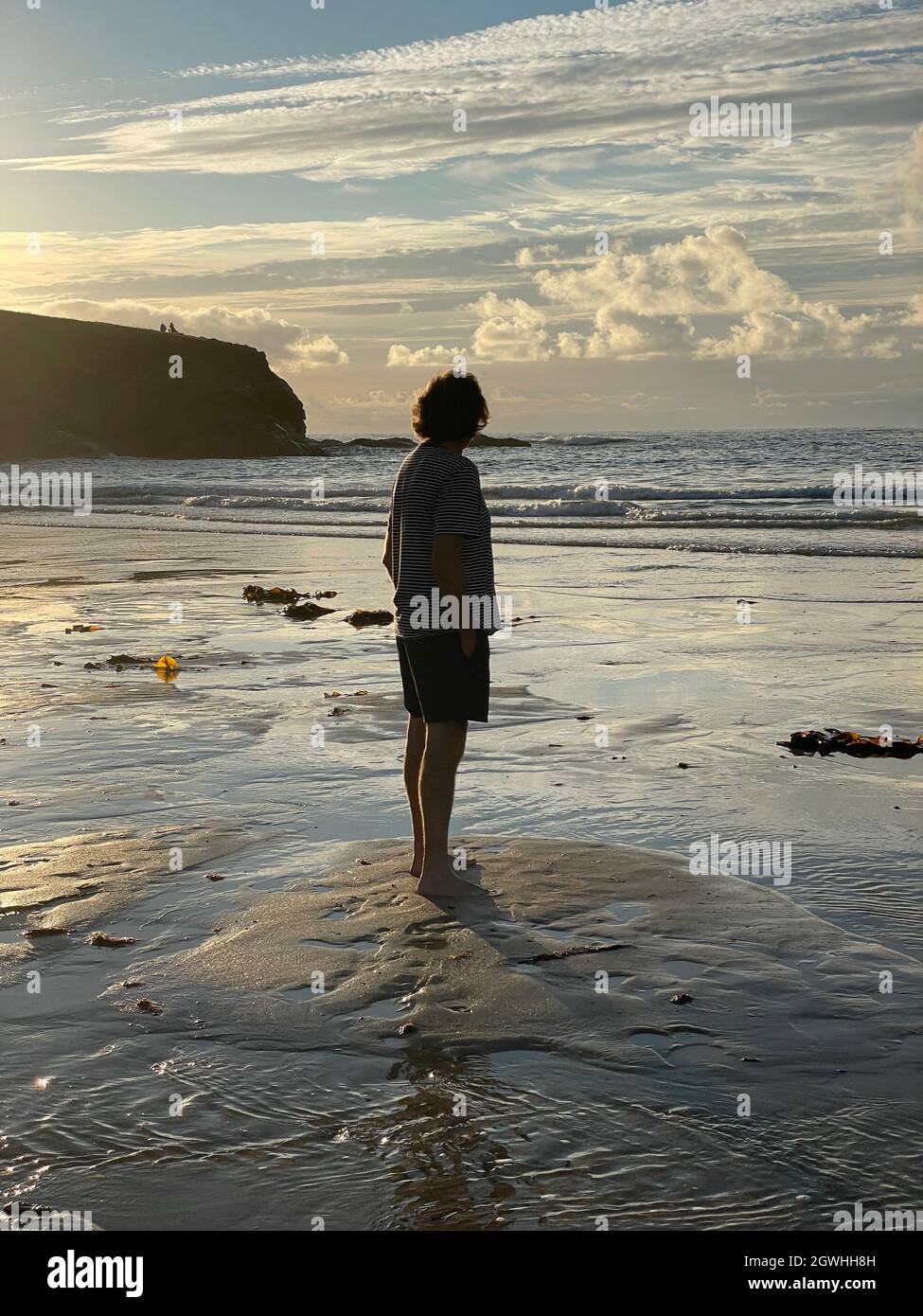 Vista posteriore a tutta lunghezza di un uomo maturo in piedi sulla spiaggia nel tardo pomeriggio mentre il sole tramonta. Guardando verso l'orizzonte indossare pantaloncini e maglietta. Foto Stock
