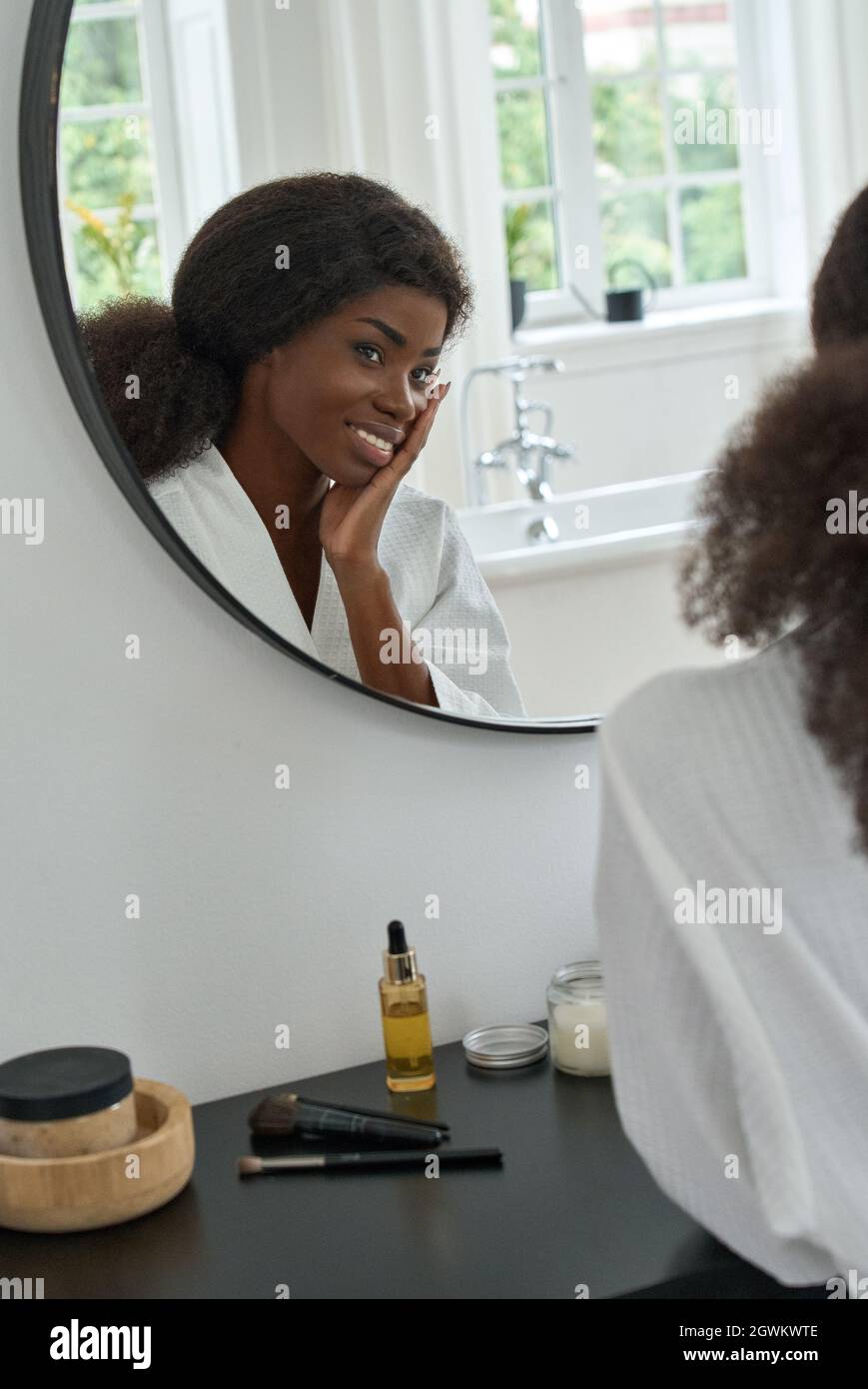 Giovane ragazza di bellezza nera che tocca il viso guardando lo specchio in bagno a casa. Foto Stock