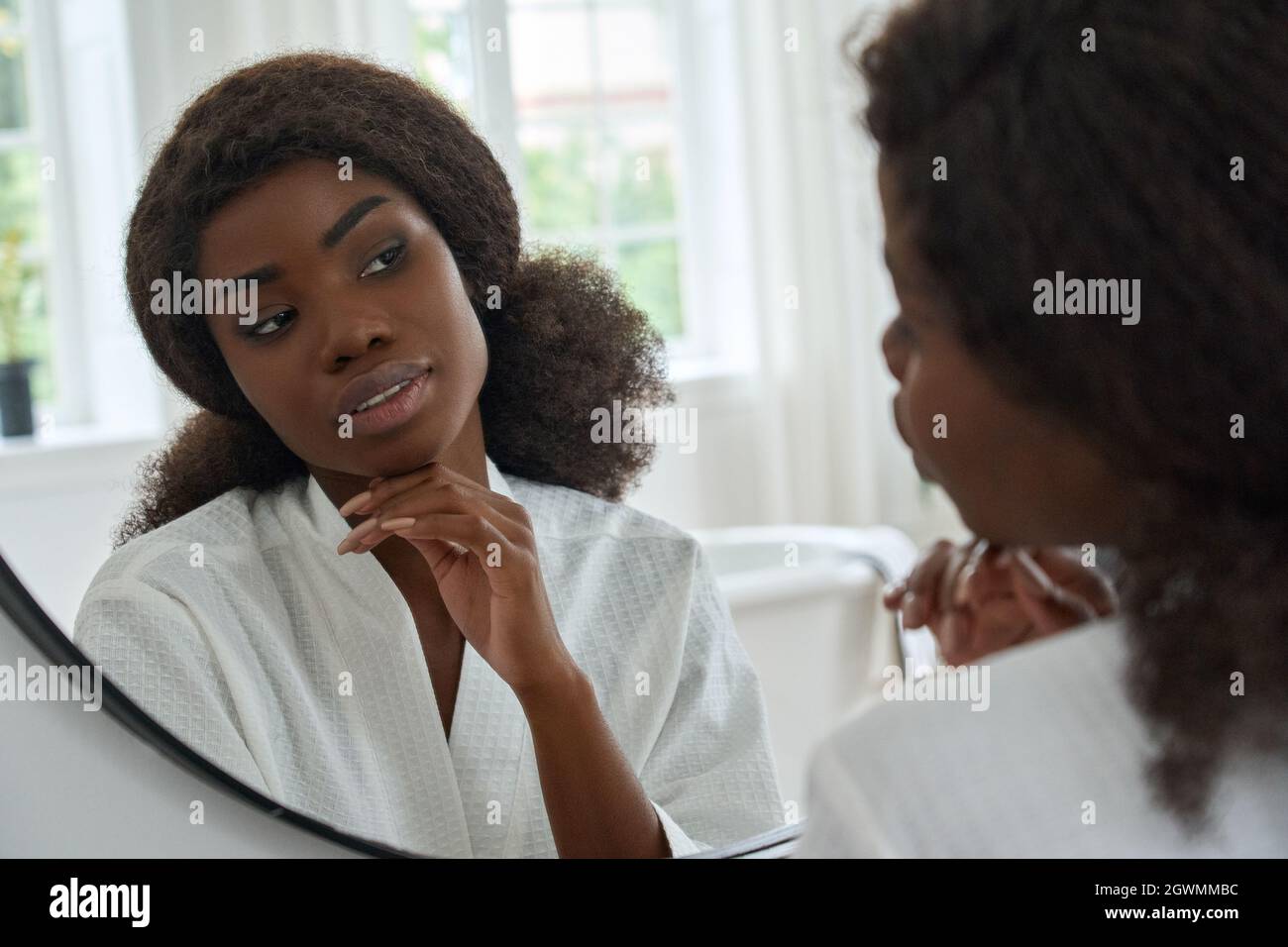 La giovane ragazza nera che tocca il viso guarda il riflesso dello specchio nella stanza da bagno a casa. Foto Stock