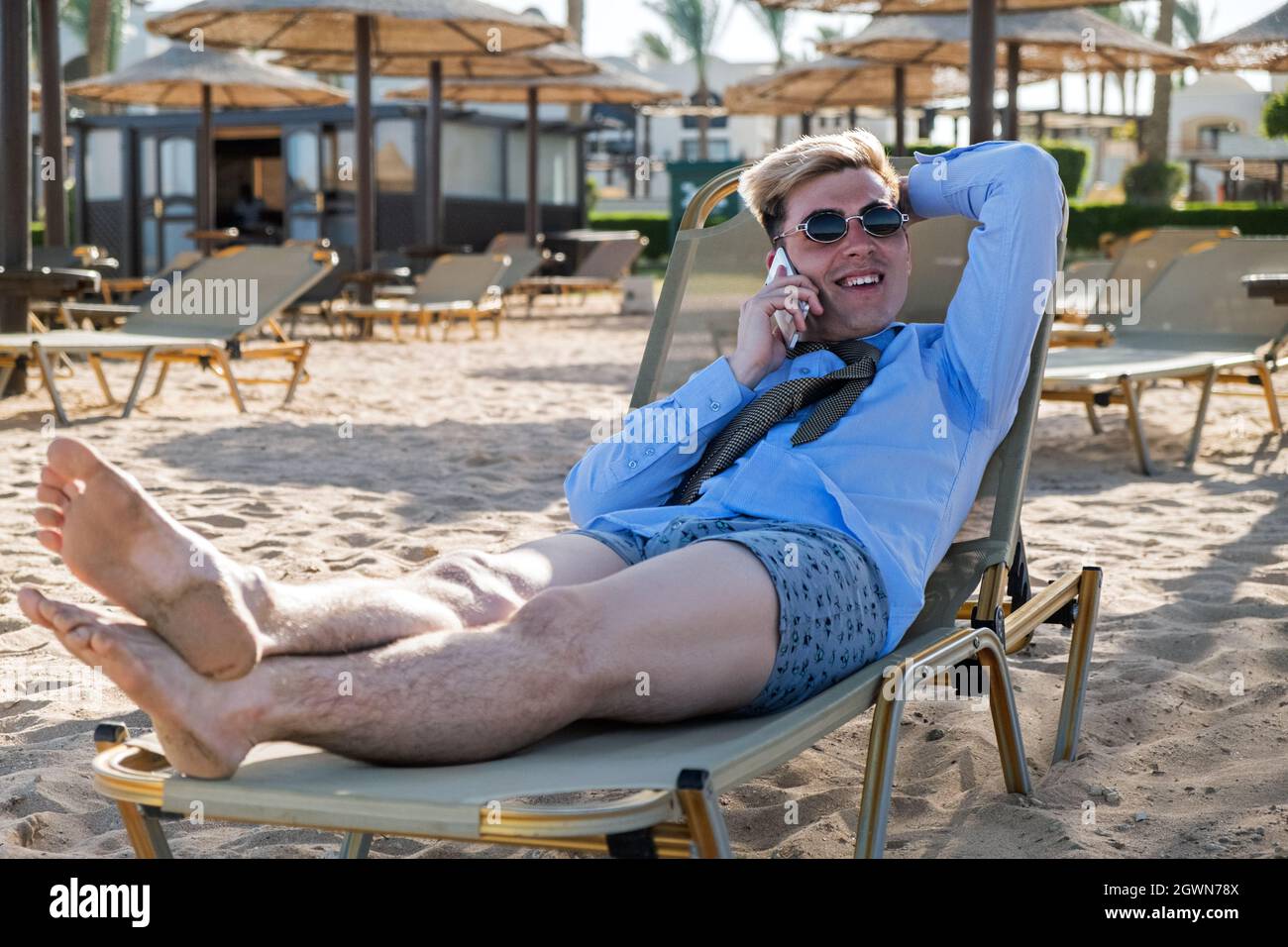 Giovane uomo d'affari felice downshifter parla al telefono sdraiato su un lettino al sole in mezzo alla spiaggia in una camicia, cravatta e boxer shorts Foto Stock