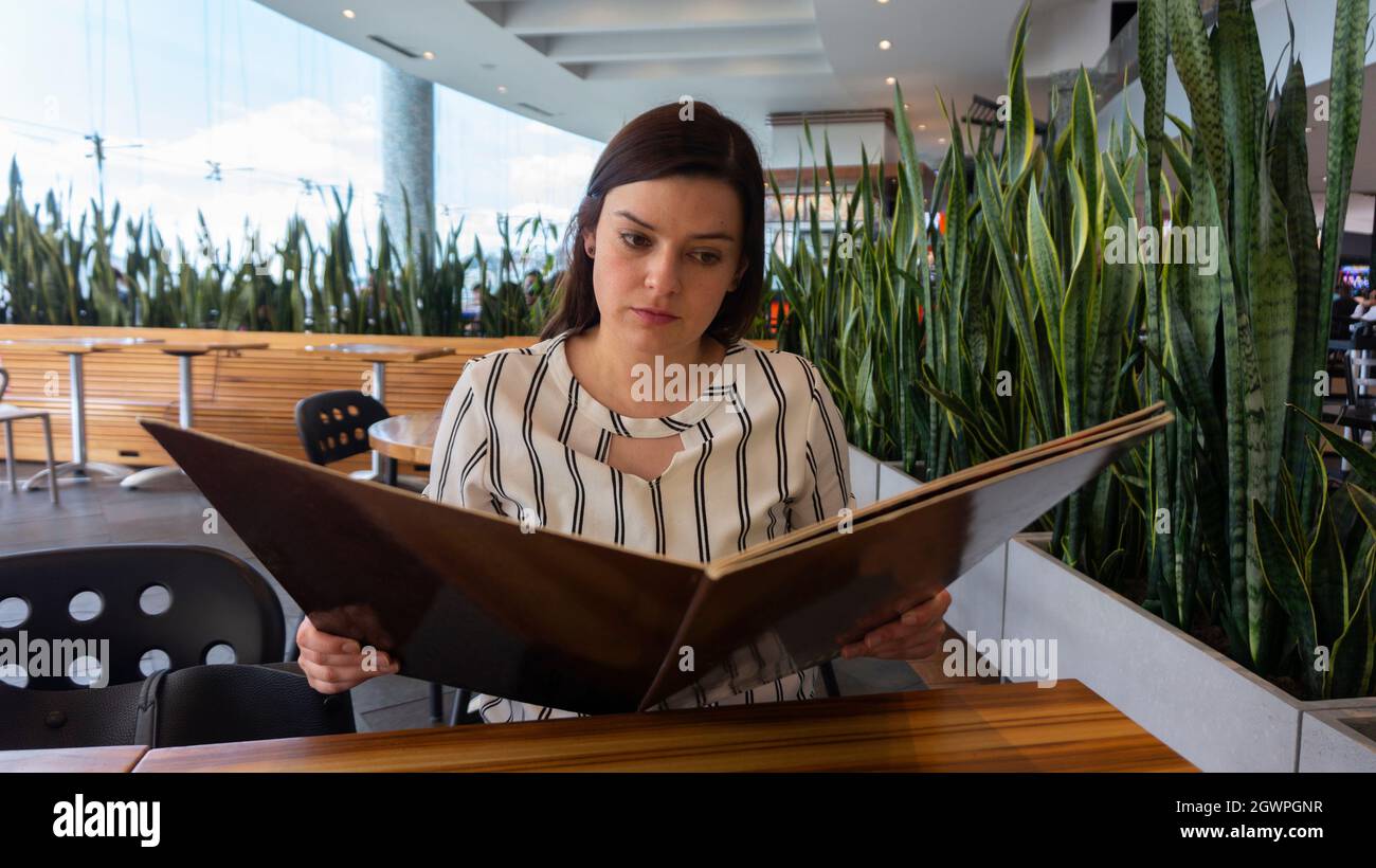 Primo piano su una bella donna ispanica che legge il menu seduto di fronte a un tavolo in un ristorante adornato di piante Foto Stock