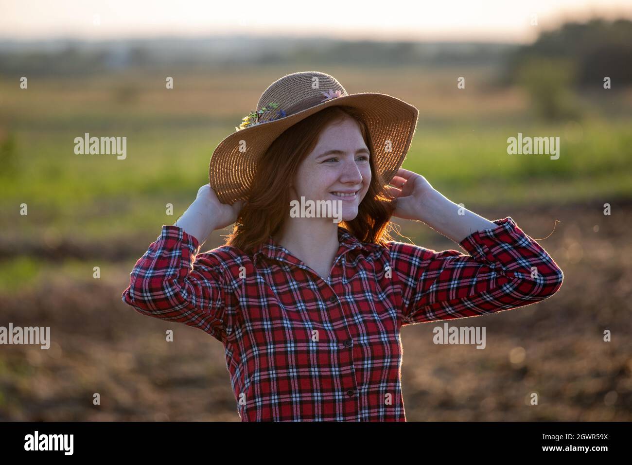 Giovane agricoltore rosso in piedi in campo sorridente. Attraente cappello da donna con entrambe le mani sulla testa al tramonto. Foto Stock