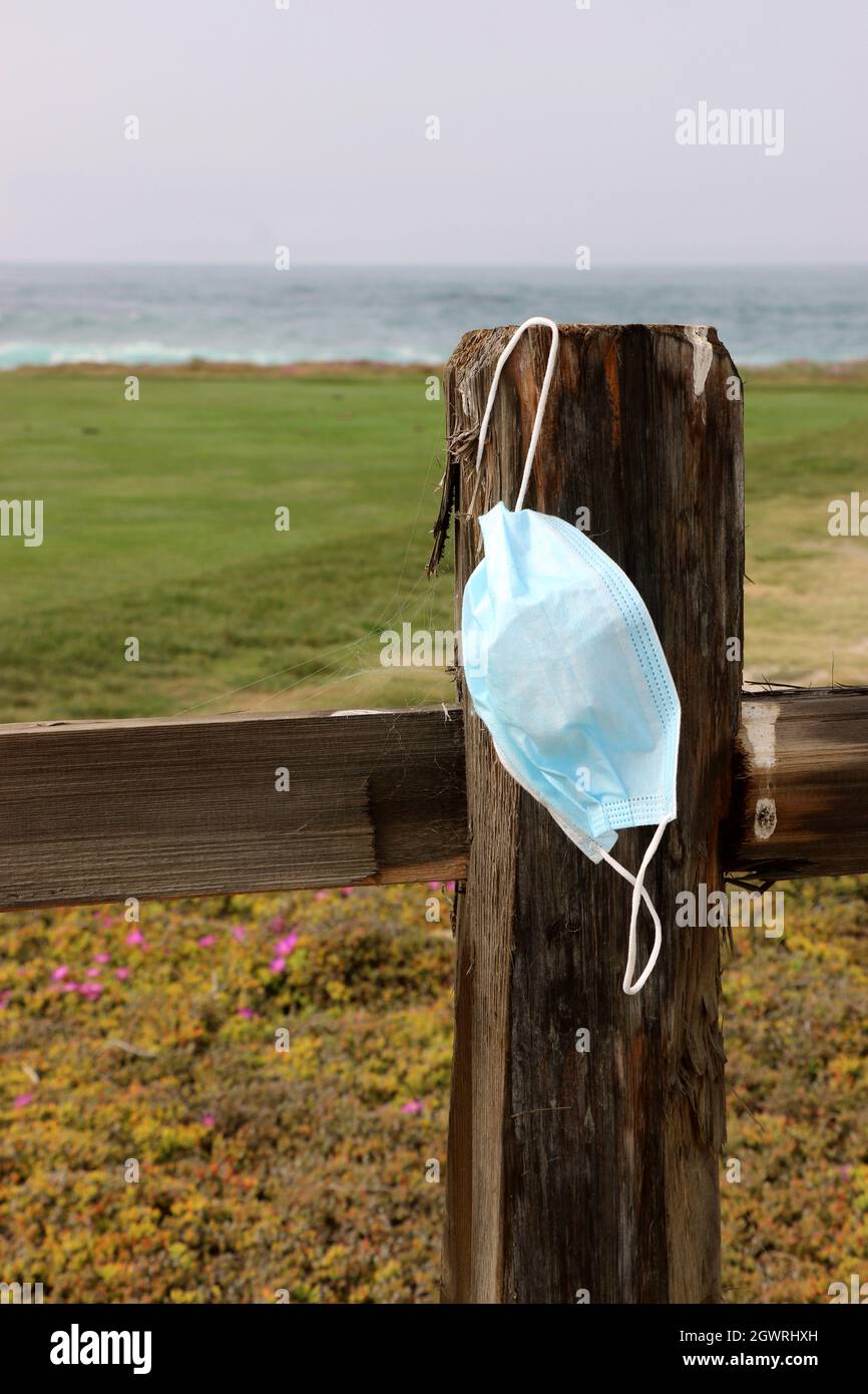 La maschera chirurgica blu scartata è appesa a un palo di recinzione resistente alle intemperie su un sentiero da spiaggia. Foto Stock