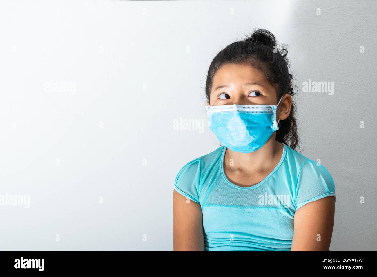 Ragazza brunetta con blusa blu e maschera chirurgica (Latina 8 anni) pensivo mentre si guarda in su preoccupato, pandemico, quarantena, covid-19. Medico concep Foto Stock