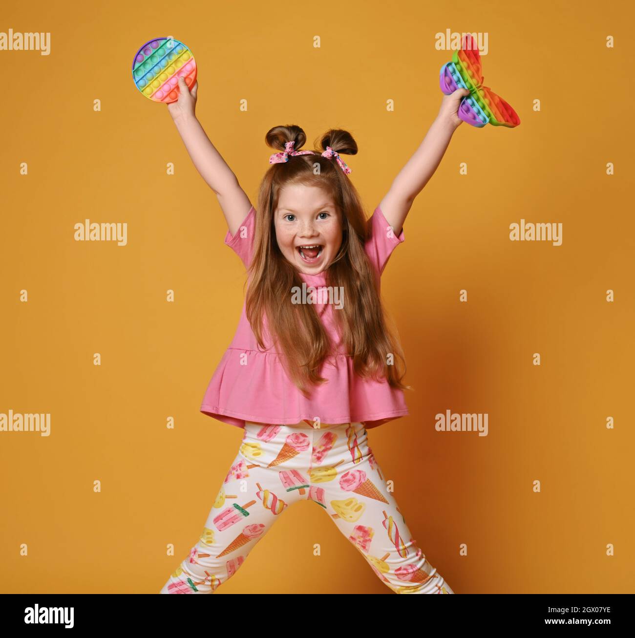 La ragazza del capretto in vestiti colorati si alza con le gambe larghe a parte, tiene due giocattoli sensoriali del colore dell'arcobaleno - schioccalo in su e grida Foto Stock