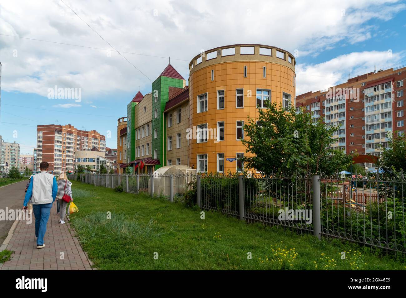 La gente cammina lungo il marciapiede passando un edificio asilo sullo sfondo di edifici residenziali in un quartiere in una giornata estiva. Foto Stock