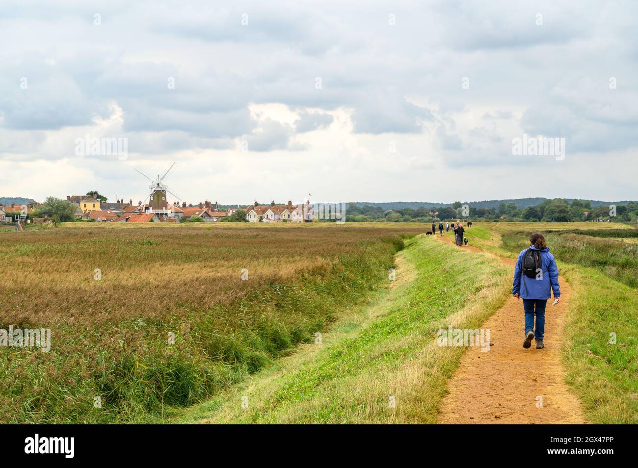 Persone che camminano sul sentiero circolare Blakeney Freshes con vista a Cley sul villaggio di mare con il suo mulino a vento, Norfolk, Inghilterra. Foto Stock