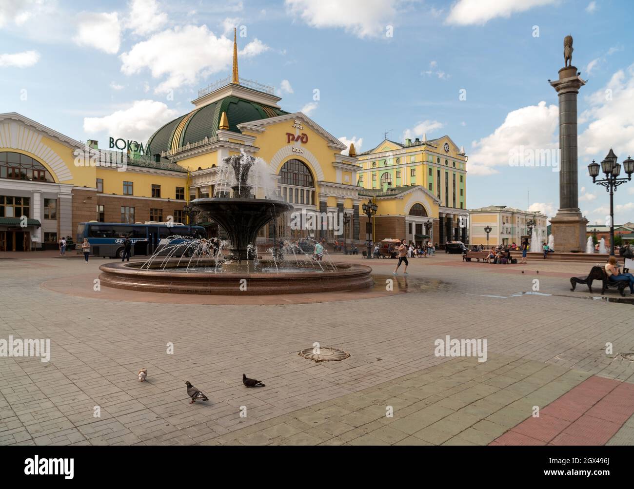 I piccioni camminano sulla piazza della stazione di fronte alla stazione ferroviaria principale della città con fontane e un pilastro con il simbolo della città su una Summe Foto Stock