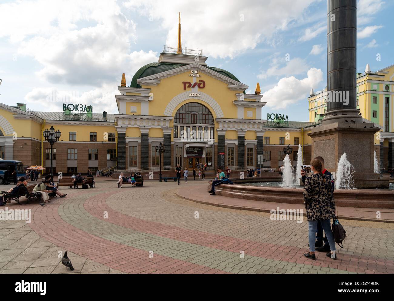 La gente riposa alla fontana sulla Piazza della Stazione ferroviaria di fronte alla stazione ferroviaria principale con il logo delle Ferrovie russe in russo sulla facciata Foto Stock