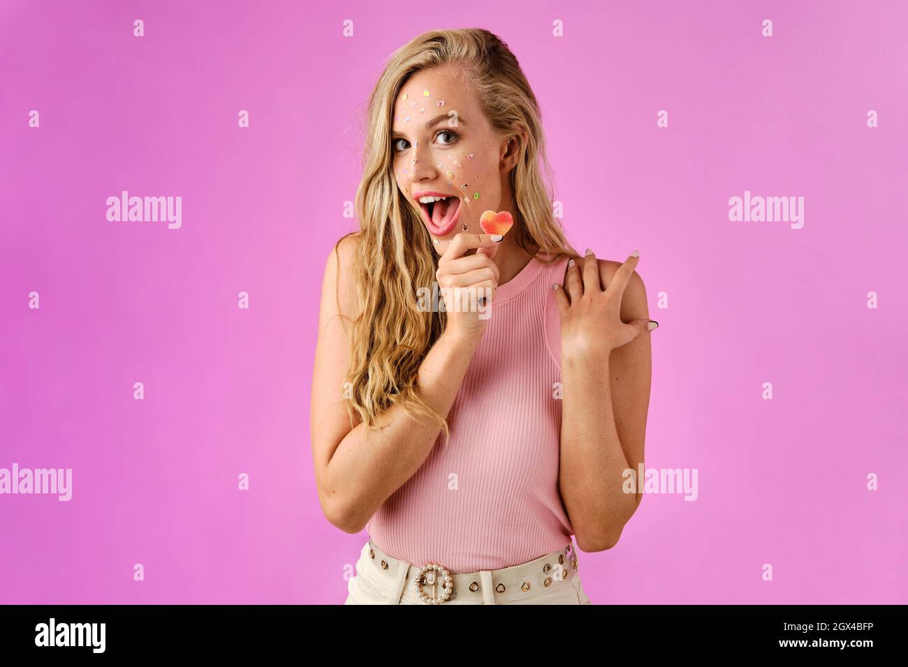 Ragazza bionda graziosa con gli adesivi sul suo viso tenendo una caramella a forma di cuore in mano su uno sfondo rosa Foto Stock