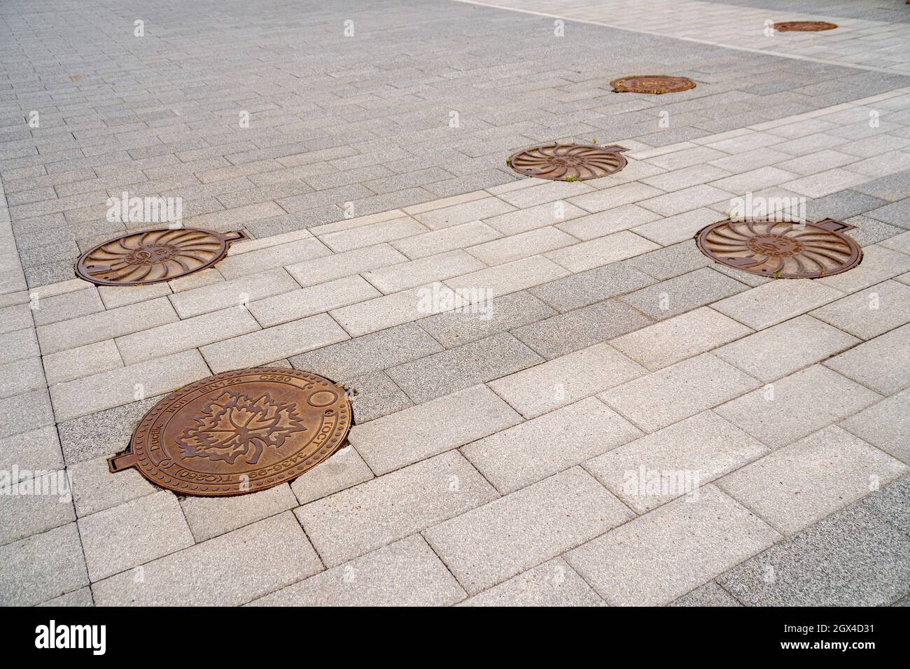 Le coperture del tombino di ghisa vintage della fogna sono situate sul marciapiede pavimentato delle mattonelle grigie di calcestruzzo. Foto Stock