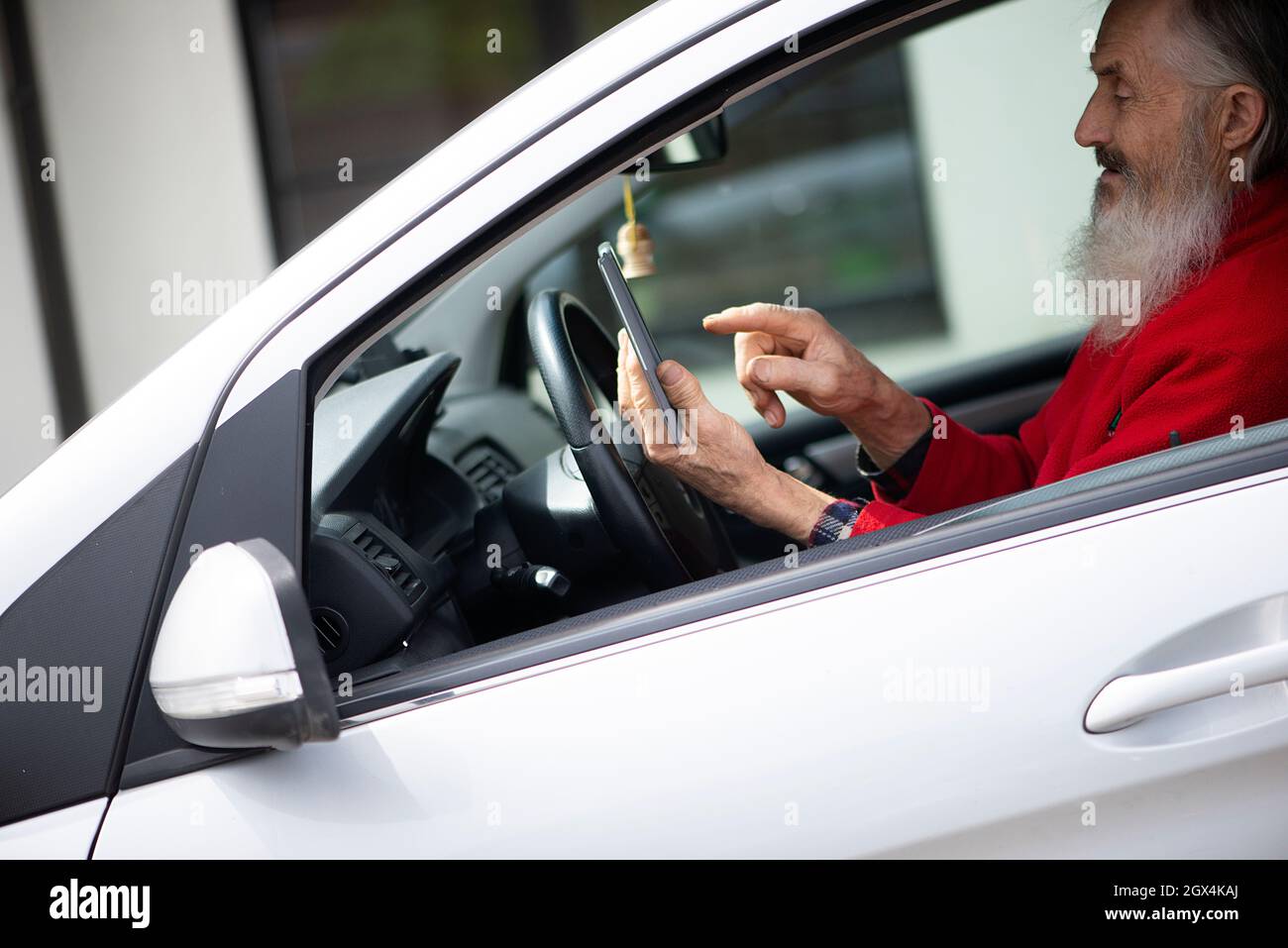 Uomo anziano bearded che tiene il telefono e che texting mentre si siede in automobile un lotto di parcheggio Foto Stock