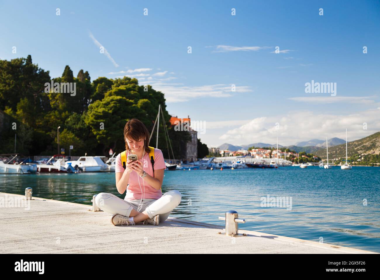 Sorridente giovane femmina turista seduto a gambe incrociate sul molo tenendo cellulare con costa croata in background. Giornata di sole a Dubrovnik, Croazia. Concetto di stile di vita di viaggio Foto Stock