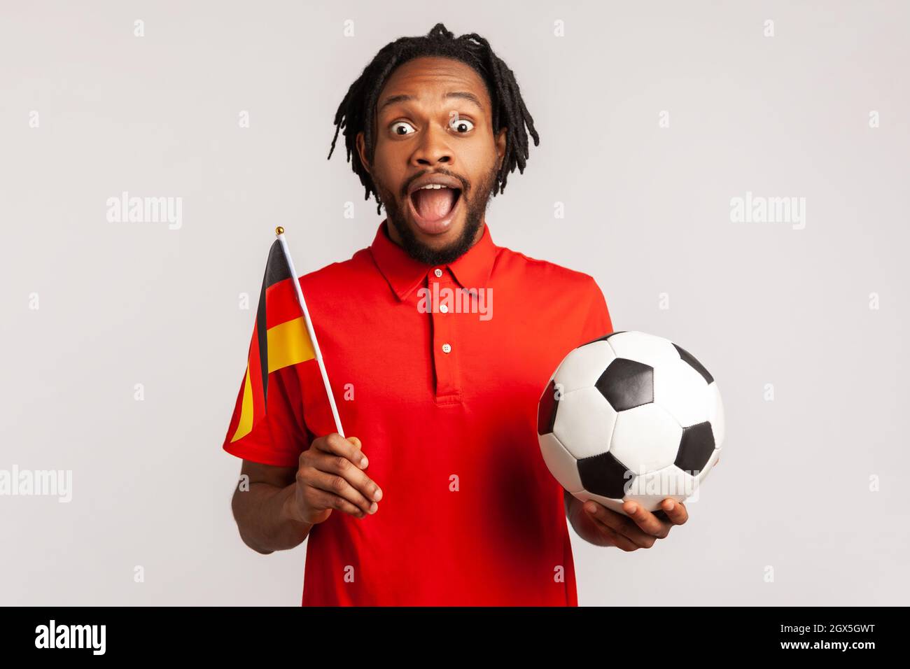 L'uomo stupito con i dreadlock che indossano una T-shirt rossa casual, che sostiene la squadra di calcio tedesca in campionato, allegria e saluto, patriottismo. Studio interno girato isolato su sfondo grigio. Foto Stock