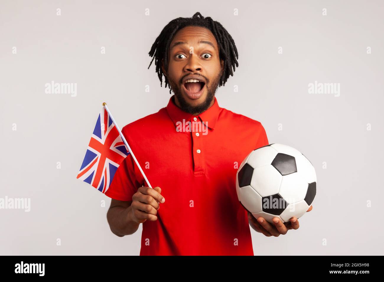 L'uomo stupito eccitato con i dreadlock che indossano una T-shirt rossa casual, che tiene la bandiera britannica e la palla di calcio in bianco e nero, la lega di calcio unita. Studio interno girato isolato su sfondo grigio. Foto Stock