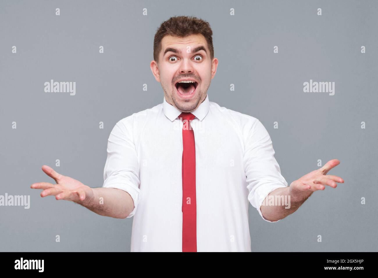Ritratto di giovane bello scioccato o sorpreso uomo in camicia bianca e cravatta in piedi con il viso scioccato, bocca aperta e braccia sollevate guardando la macchina fotografica. Interno isolato su sfondo grigio. Foto Stock