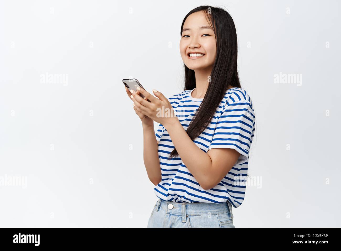 Ritratto di ragazza asiatica con smartphone e sorridente alla fotocamera, concetto di tecnologia cellulare e applicazioni mobili, sfondo bianco Foto Stock