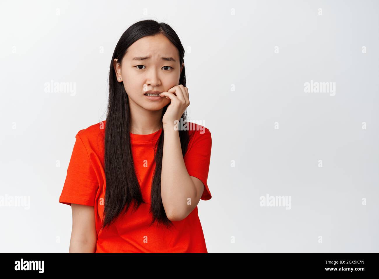 Spaventato ragazza asiatica che morde le dita e guarda la macchina fotografica insicura, ansiosa di qualcosa, si sente preoccupato, in piedi su sfondo bianco Foto Stock