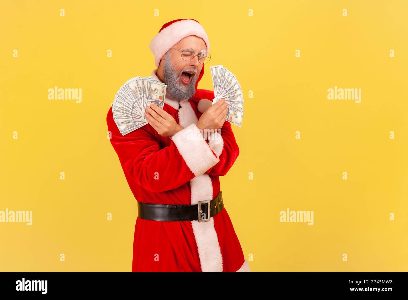 Uomo anziano felice eccitato con barba grigia che indossa il costume di babbo natale in piedi con due fan di banconote, guardando la macchina fotografica e l'argentatura. Studio interno girato isolato su sfondo giallo. Foto Stock