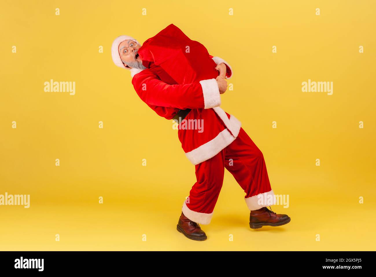 Uomo anziano a tutta lunghezza con barba grigia che indossa il costume di babbo natale e che trasporta una grande borsa pesante con regali di Natale, congratulandosi con Capodanno. Studio interno girato isolato su sfondo giallo. Foto Stock