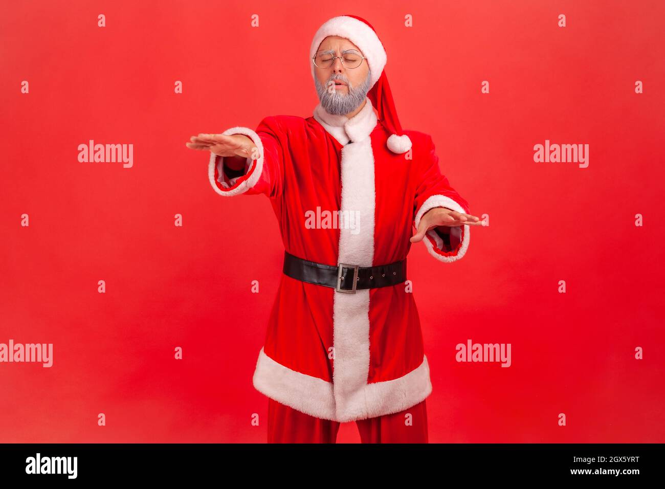 Problemi di visione. Ritratto di cieco disorientato solitario anziano in costume di babbo natale a piedi con occhi chiusi, alzando le mani per cercare la strada persa. Studio interno girato isolato su sfondo rosso. Foto Stock