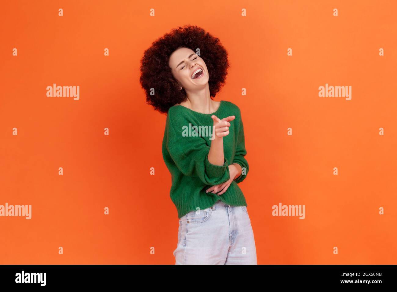 Donna con acconciatura afro che indossa un maglione verde che tiene la mano sullo stomaco e ride fuori forte, puntando il dito alla fotocamera, indicando ridicolo idiota. Studio interno girato isolato su sfondo arancione. Foto Stock